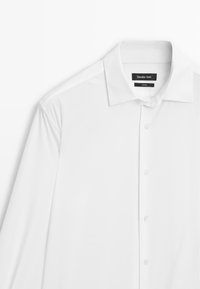 Biała koszula z kołnierzykiem, wykonana z gładkiego materiału. Posiada czarną metkę z logo poniżej kołnierzyka oraz przezroczyste guziki.