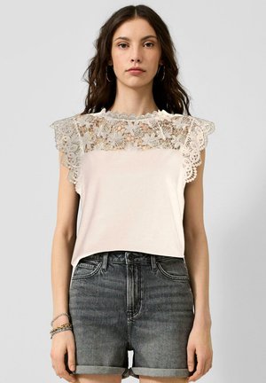 Femme aux longs cheveux foncés portant un haut sans manches blanc en dentelle crochetée et un short en jean foncé taille haute retroussé, debout devant un fond uni.