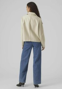 Krämfärgad, strukturerad jacka med krage och lagerdesign bak, parat med blå vida jeans och svarta hälstövlar.