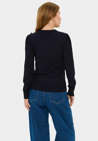 Granatowy sweter z dzianiny z bufkami na rękawach, ściągaczami przy mankietach i na dole oraz dekoracyjnymi guzikami na ramieniu. W połączeniu z dżinsami o szerokich nogawkach.