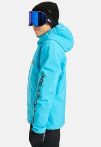 Veste imperméable bleue avec capuche, arborant un logo noir sur la poitrine et une inscription noire le long de la manche. Portée avec un bonnet en tricot noir et des lunettes de protection.