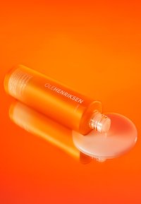 OLEHENRIKSEN leuchtend orangefarbene Glasflasche mit einer weißen Pumpe, die geprägten Text zeigt. Ein klarer runder Deckel in der Nähe enthüllt eine flüssige Essenz.