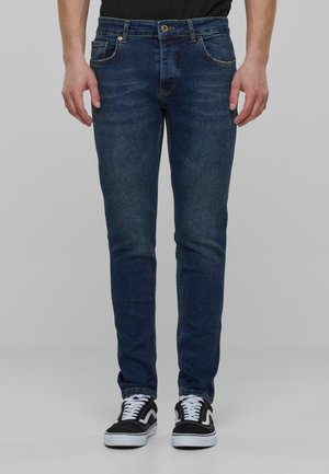 Jeans de mezclilla azul oscuro con ajuste slim, que cuentan con cinco bolsillos, remaches de cobre y un ligero desvanecimiento en los muslos. Combinados con zapatillas negras.