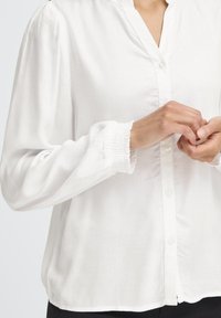 Blusa bianca con colletto alla mandarino, chiusura a bottoni e polsini arricciati. Realizzata in tessuto liscio, presenta una vestibilità comoda e una leggera lucentezza.