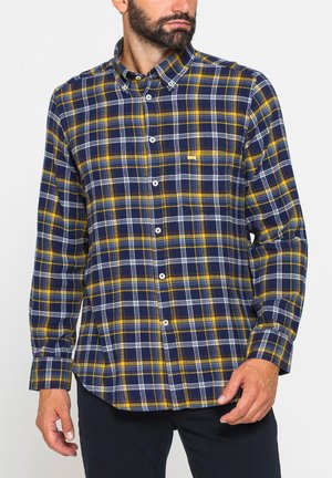 Uomo che indossa una camicia a maniche lunghe con motivo a quadri blu, giallo e bianco, in piedi davanti a uno sfondo semplice.
