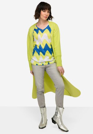 Cárdigan largo verde sobre una blusa con patrón de zigzag azul y amarillo, combinado con pantalones grises y botas de tobillo plateadas. Texturas suaves en toda la composición.