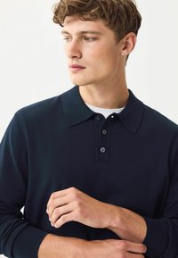 Polo de culoare bleu marin, cu mânecă lungă, fabricat dintr-un material texturat, cu guler clasic și închidere frontală cu trei nasturi; modelul poza cu mâinile aproape de piept.
