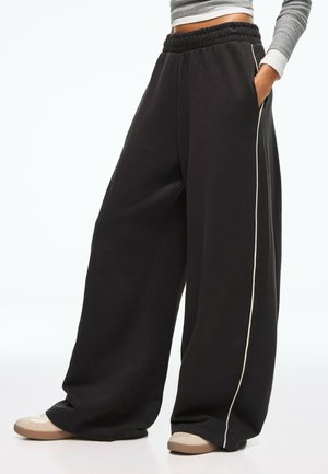 Osoba nosząca czarne spodnie z szerokimi nogawkami z białymi paskami po bokach i beżowe buty, stojąca z jedną ręką w kieszeni.