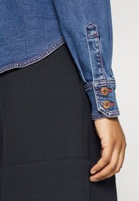 Chemise en jean avec tissu bleu foncé, coutures rouges et poignets boutonnés. Matière textile noire visible en dessous, avec un détail de poche.