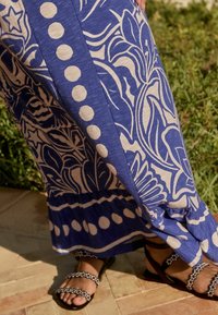 Personne portant un pantalon large à motifs bleus et blancs et des sandales noires avec des perles décoratives, debout sur un sol carrelé près de la verdure.