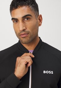 Svart zip-up jacka med uppstående krage och vit "BOSS"-logotyp. Slät tyg, figurnära passform och en kontrasterande lila innerfoder.