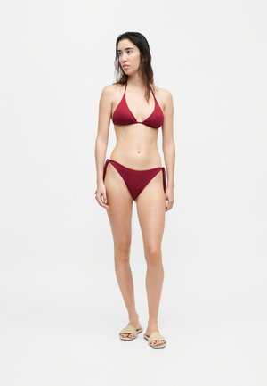 Žena s mokrými vlasmi, oblečená v červených bikinách s halterom a béžových pletených sandáloch, stojaca pred jednofarebným svetlým pozadím.