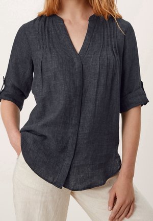 Femme portant un chemisier à manches longues gris foncé retroussées et un pantalon beige clair, une main dans la poche, debout devant un fond neutre.