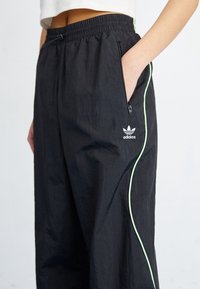 Persona vistiendo pantalones de chándal negros de Adidas con bandas laterales de color verde claro y una mano en el bolsillo con cremallera, mostrado desde la cintura hasta la mitad del muslo.
