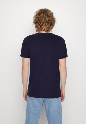 Person med krøllet blondt hår står med ryggen mod, iført en ensfarvet mørkeblå kortærmet t-shirt og lyseblå jeans mod en hvid baggrund.