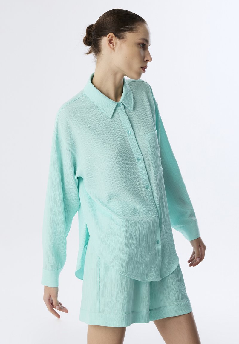 Camisa azul turquesa clara de gran tamaño, hecha de tela texturizada, con cierre de botones en la parte delantera y un solo bolsillo en el pecho, combinada con pantalones cortos texturizados a juego.