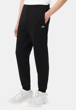 Pantaloni della tuta neri con vita elastica, tasche laterali e un discreto logo verde Lacoste. Realizzati in materiale morbido, con una vestibilità affusolata verso la caviglia.