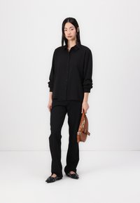 Black button-up shirt with long sleeves, paired with black trousers and black flat shoes, holding a brown bag with a shoulder strap.