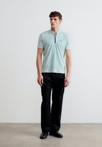 Νεαρός άνδρας στέκεται φορώντας φωτεινό γαλάζιο πόλο με κοντά μανίκια και μαύρο παντελόνι με μαύρα παπούτσια σε απλό φόντο.