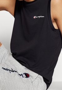 Svart ärmlös linne med en vit "Champion"-logotyp. Bärs tillsammans med grå shorts där "Champion"-namnet är broderat i marinblått.