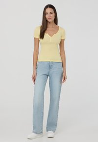 Top giallo a maniche corte con scollatura a barchetta e tre bottoni, abbinato a jeans a gamba larga azzurri e sneakers bianche.