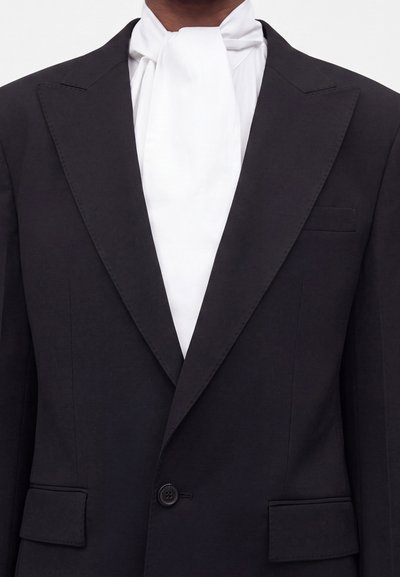 Homme portant une veste de smoking noire avec de larges revers et une cravate blanche de style lavallière sur une chemise blanche.