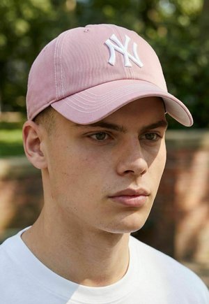 Junger Mann, der eine hellrosa Baseballkappe mit weißem "NY"-Logo trägt, im Freien vor einem unscharfen Hintergrund aus Grünpflanzen und einer Backsteinmauer.
