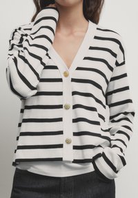 Striped cardigan featuring horizontal black and white stripes, ribbed texture, gold buttons, and a V-neck design. Tailored fit with long sleeves.