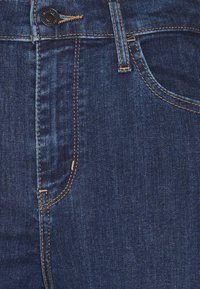 Gros plan d’un jean en denim bleu foncé montrant le bouton, la zone de la fermeture éclair, la passants de ceinture et la poche avant avec une couture orange contrastée.