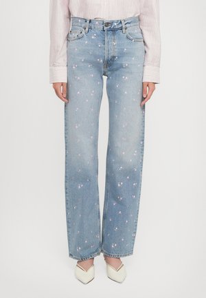 Woman wearing light blue high-waist jeans with small pink floral spots, a striped shirt, and white pointed-toe shoes on a plain background.