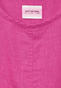 Tissu en lin rose avec un col rond. Une étiquette indique "JUSTE NATUREL POUR LES AMATEURS DE QUALITÉ" en texte noir sur un fond rose clair.