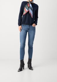 Pullover blu, jeans skinny blu e stivaletti neri alla caviglia con punte in argento. Donna accessoriata con una sciarpa a righe colorate attorno al collo.