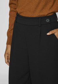 Pantalon noir à coupe ajustée, présentant une texture lisse, une taille plate et une fermeture à bouton. Détails de pinces à l'avant visibles.