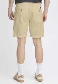 Pantaloni corti in cotone beige con risvolti. Due tasche posteriori con bottoni marroni. Indossati con calze bianche e sneakers beige con accenti dorati.