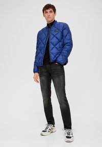 Modrá prešívaná bomber bunda s vysokým golierom, so zipsom vpred, bočnými vreckami a logom, skombinovaná s tmavými džínsami a teniskami.