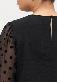 Zwarte top van textuurstof, met doorzichtige zwarte pofmouwen versierd met stippen en een sleutelgatopening aan de achterkant.