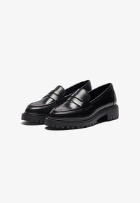 Pantofi loafer din piele neagră cu o talpă groasă neagră, având un finisaj neted, o curea largă în față și un design cu vârf rotunjit.