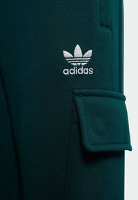 Pantalons de jogging verts en tissu doux, dotés d'une poche à rabat sur le côté et d'un logo brodé blanc avec trois bandes.