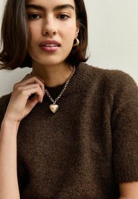 Pull en tricot marron à manches courtes, arborant un pendentif en forme de cœur doré sur une chaîne épaise. Texture lisse et accents métalliques visibles.
