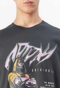 Person wearing dark gray t-shirt with colorful graphic of a hooded, metallic skeletal figure and stylized text reading "Adidas Originals".