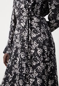 Robe noire à motif floral blanc, manches longues et ceinture en tissu fin nouée à la taille, portée par une personne à la peau foncée.
