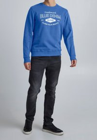 Sudadera azul con cuello redondo, texto gráfico en blanco, combinada con jeans negros y zapatillas negras. Textura de tela suave, ajuste casual.