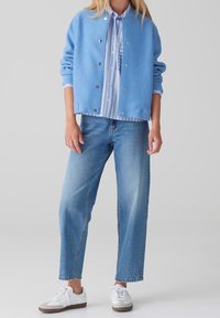 Femme portant une veste bleu clair boutonnée sur une chemise rayée, un jean bleu et des baskets blanches, debout devant un fond gris.