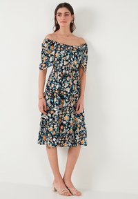 Off-shoulder cvjetna haljina s tamnoplavom podlogom i narančastim te tirkiznim cvjetovima, uskim korzetom i lepršavom suknjom s višeslojnim detaljima.