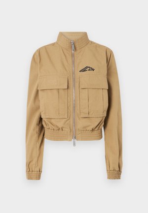 Veste beige cintrée avec un col haut, fermeture éclair à l'avant et deux grandes poches poitrine, dotée de poignets élastiques et d'un logo sur le côté gauche.