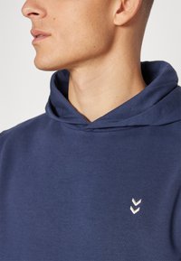 Felpa navy in morbido cotone, caratterizzata da un cappuccio prominente, un collo a coste e un piccolo logo a chevron bianco sul davanti.