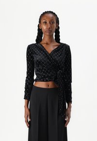 Μαύρη βελούδινη μπλούζα με μακριά μανίκια, με διαγώνιο μοτίβο από ασημένια διαμάντια και με κόμπο μπροστά. Συνδυάζεται με παντελόνι με φαρδιές γάμπες.