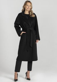 Czarny dwurzędowy płaszcz z wiązaniem w talii i szerokimi rękawami, wykonany z teksturowanej mieszanki wełny, w połączeniu z czarnymi dopasowanymi spodniami i szpilkami.