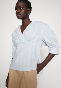 Lys blåstripet bluse med v-hals, rynkede detaljer i midjen, og puffede ærmer. Kombinert med beige bukser.