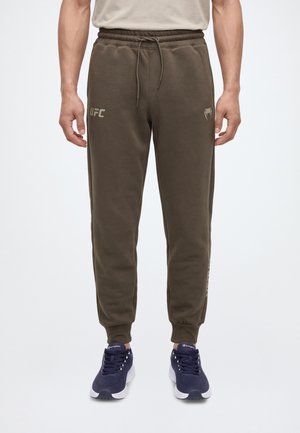Modelo con pantalones de chándal UFC de color verde oliva, con cordón en la cintura y puños en los tobillos, combinados con zapatillas deportivas azul marino sobre un fondo blanco.